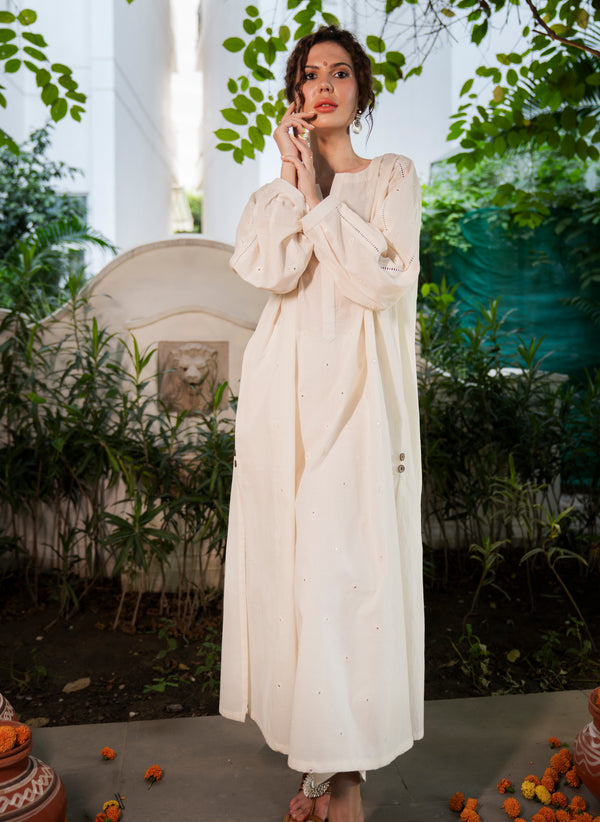 Woman in a long, light-colored dress standing outdoors with greenery in the background